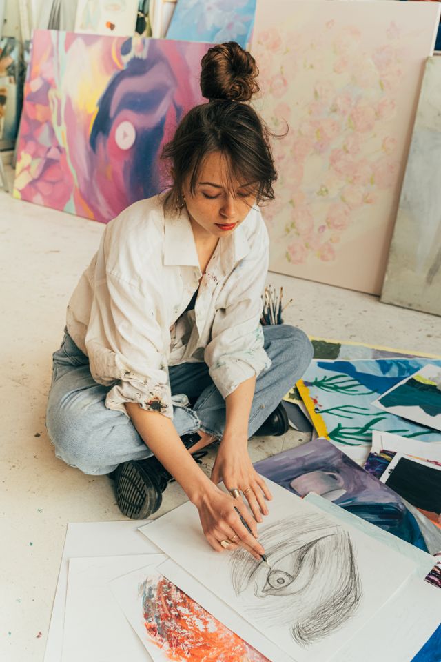 Artist sitting on floor sketching in a vibrant art studio filled with colorful paintings.