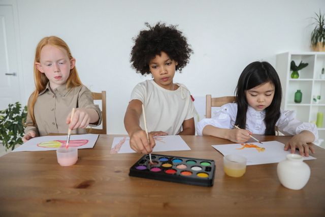 Focused cute multiracial preteen female friends sitting at wooden table and dipping paintbrushes into water and aquarelle while drawing together at home